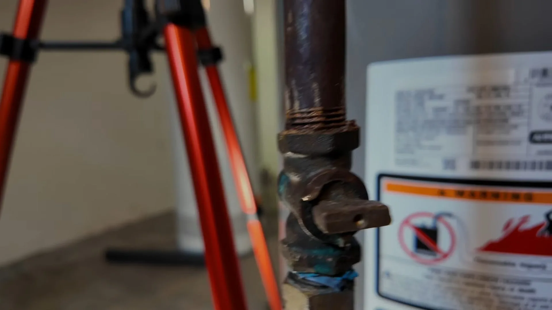 Gas water heater with a brass shutoff valve and a warning label, and a red tripod stand behind it