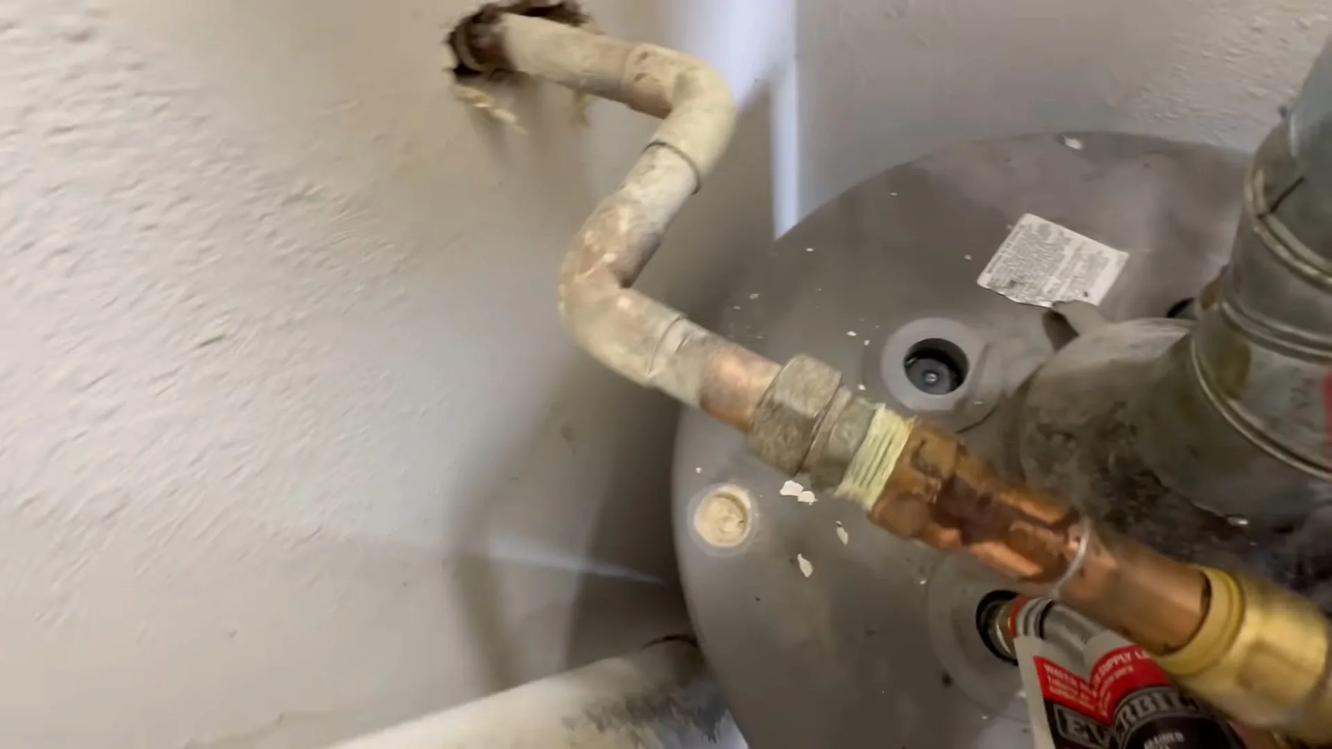 Top of a tank water heater with a flexible connector and brass fittings leading into the wall
